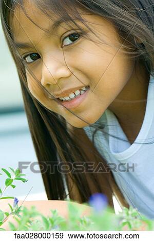 Stock Photo - Little girl leaning over flowering plant, hair tousled by breeze, smiling at camera. Fotosearch
