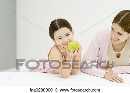 Little girl sitting at table with mother, hand under chin, holding apple, smiling at camera View Large Photo Image Stock Image - Little girl sitting at table with mother, hand under chin, holding apple, smiling at camera. Fotosearch