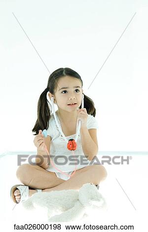 Stock Photo - Little girl sitting next to teddy bear, listening to toy stethoscope. Fotosearch