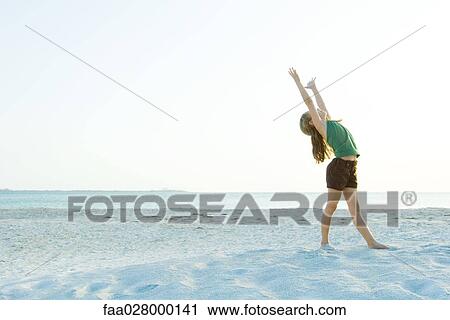 Stock Image - Little girl standing on the beach with arms raised, head back, full length. Fotosearch