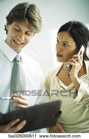 Male and female colleagues standing side by side, woman using cell phone, man looking at binder View Large Photo Image Stock Image - Male and female colleagues standing side by side, woman using cell phone, man looking at binder. Fotosearch