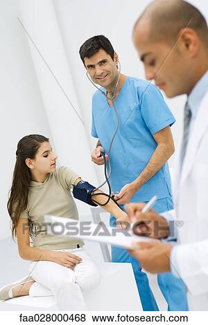 Male nurse measuring young patient's blood pressure, smiling at camera, doctor writing on clipboard in foreground View Large Photo Image Stock Photo - Male nurse measuring young patient's blood pressure, smiling at camera, doctor writing on clipboard in foreground. Fotosearch