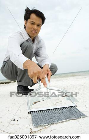 Man crouching on beach, sweeping sand into dustpan, looking at camera, low angle view View Large Photo Image Stock Photo - Man crouching on beach, sweeping sand into dustpan, looking at camera, low angle view. Fotosearch