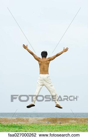 Stock Image - Man jumping in the air at the beach, arms raised, rear view. Fotosearch