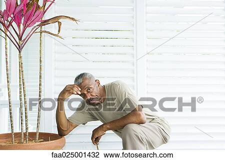 Stock Image - Man kneeling beside potted plant, holding head, looking at camera. Fotosearch