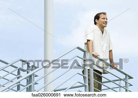 Stock Image - Man leaning against railing, looking into distance, low angle view. Fotosearch