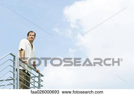 Stock Image - Man leaning against railing, looking into distance, low angle view. Fotosearch