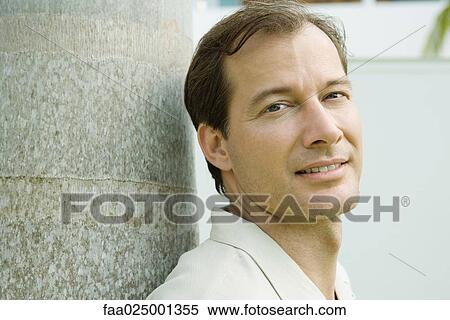 Stock Photography - Man leaning against tree trunk, smiling at camera, close-up. Fotosearch