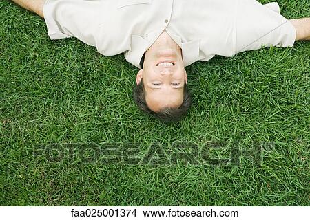 Picture - Man lying upside down in grass, smiling at camera, cropped high angle view. Fotosearch