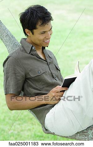 Stock Image - Man resting on tree branch, holding book. Fotosearch