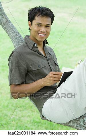 Man resting on tree branch, holding book, smiling at camera View Large Photo Image Picture - Man resting on tree branch, holding book, smiling at camera. Fotosearch