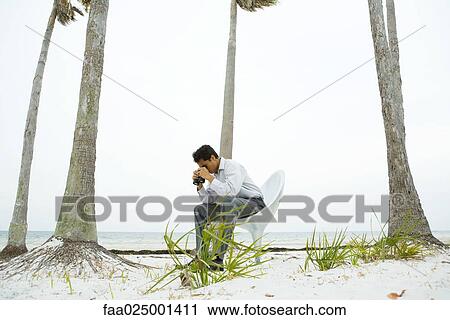 Stock Image - Man sitting in chair at the beach, looking at ground through binoculars . Fotosearch