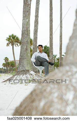 Stock Photo - Man sitting in chair at the beach, looking at camera, low angle view. Fotosearch