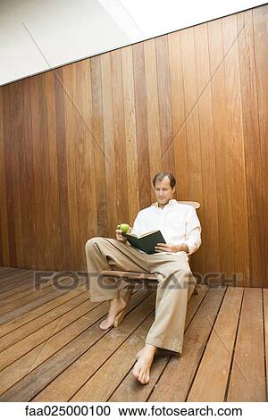 Man sitting in lounge chair reading book, apple in hand View Large Photo Image Stock Image - Man sitting in lounge chair reading book, apple in hand. Fotosearch