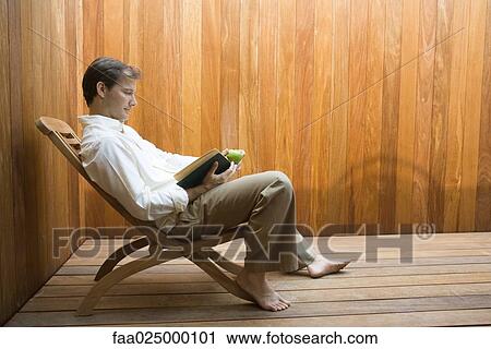 Stock Image - Man sitting in lounge chair reading book, apple in hand, side view. Fotosearch