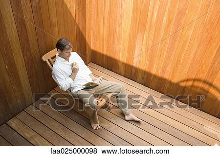 Man sitting in lounge chair reading book, apple in hand, high angle view View Large Photo Image Stock Photo - Man sitting in lounge chair reading book, apple in hand, high angle view. Fotosearch