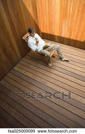 Man sitting in lounge chair reading book, apple in hand, high angle view View Large Photo Image Stock Photo - Man sitting in lounge chair reading book, apple in hand, high angle view. Fotosearch