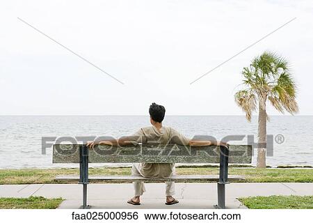 Stock Photograph - Man sitting on bench, facing ocean, rear view. Fotosearch
