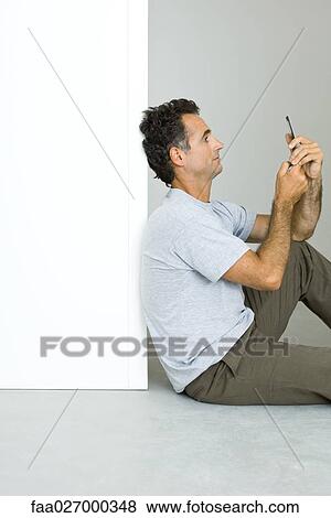 Stock Photo - Man sitting on the ground, looking at cell phone, side view. Fotosearch