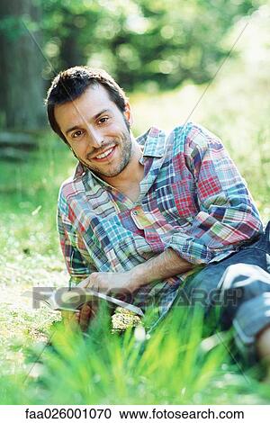 Man sitting on the ground outdoors, holding book, smiling at camera View Large Photo Image Stock Image - Man sitting on the ground outdoors, holding book, smiling at camera. Fotosearch