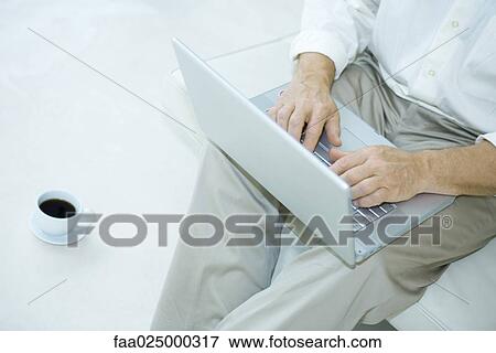 Stock Photo - Man sitting using laptop computer, cup of coffee beside him, cropped view. Fotosearch