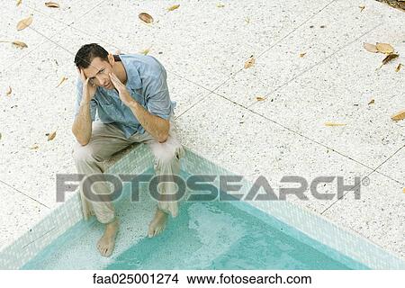 Man sitting with feet in pool, holding head, high angle view View Large Photo Image Picture - Man sitting with feet in pool, holding head, high angle view. Fotosearch