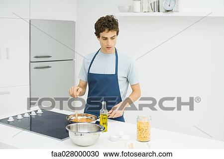 Stock Image - Man standing at stove, cooking, looking down at recipe. Fotosearch