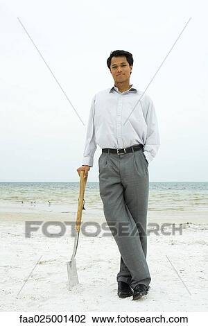 Stock Image - Man standing at the beach, leaning against shovel, smiling at camera. Fotosearch