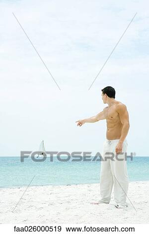 Stock Photo - Man standing at the beach, pointing at sailboat in the distance, side view. Fotosearch