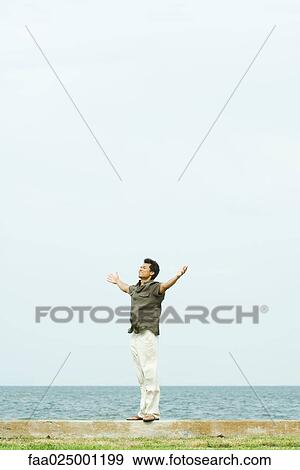 Man standing by sea with arms out and eyes closed, full length View Large Photo Image Stock Photo - Man standing by sea with arms out and eyes closed, full length. Fotosearch