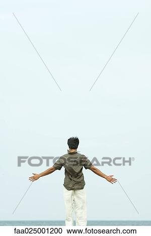 Stock Image - Man standing by sea with arms out, rear view. Fotosearch