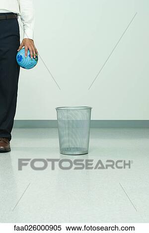Stock Photography - Man standing next to garbage can, holding globe in hand, cropped view. Fotosearch