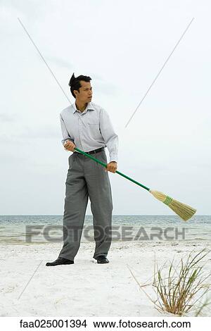 Picture - Man standing on beach sweeping with broom, looking away. Fotosearch