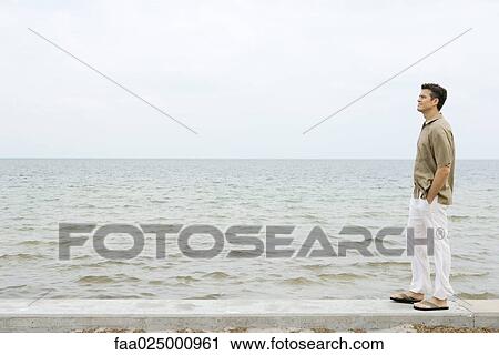 Man standing on low wall by ocean, hands in pockets, full length View Large Photo Image Stock Image - Man standing on low wall by ocean, hands in pockets, full length. Fotosearch