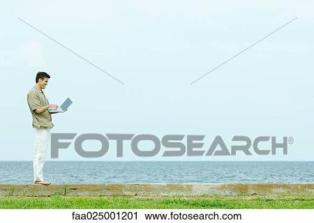 Man standing on low wall using laptop, body of water in background, full length View Large Photo Image Stock Image - Man standing on low wall using laptop, body of water in background, full length. Fotosearch