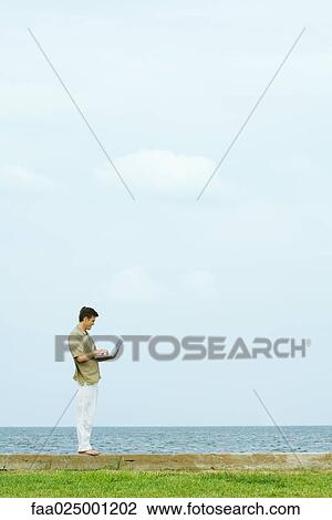 Man standing on low wall using laptop, body of water in background, full length View Large Photo Image Stock Image - Man standing on low wall using laptop, body of water in background, full length. Fotosearch