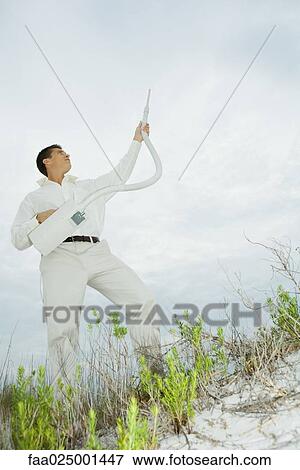 Stock Photo - Man standing on sand dune, holding up vacuum cleaner, looking up. Fotosearch