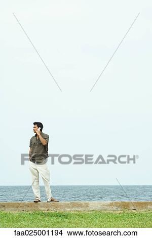 Man standing, using cell phone, body of water in background View Large Photo Image Picture - Man standing, using cell phone, body of water in background. Fotosearch