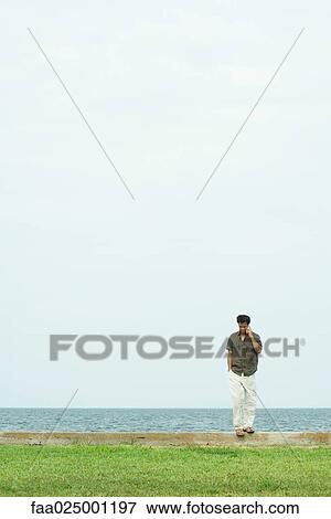 Stock Photo - Man standing, using cell phone, body of water in background. Fotosearch