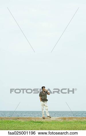 Man standing, using cell phone, body of water in background View Large Photo Image Stock Photograph - Man standing, using cell phone, body of water in background. Fotosearch