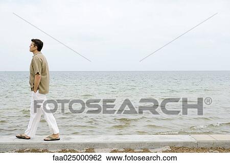 Man walking along low wall by ocean, hands in pockets, full length View Large Photo Image Stock Image - Man walking along low wall by ocean, hands in pockets, full length. Fotosearch