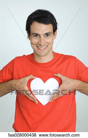 Man wearing tee-shirt with heart symbol, holding hands around heart, smiling at camera View Large Photo Image Stock Photo - Man wearing tee-shirt with heart symbol, holding hands around heart, smiling at camera. Fotosearch
