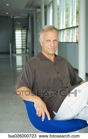 Stock Image - Mature man sitting in chair in hallway, smiling at camera, portrait. Fotosearch