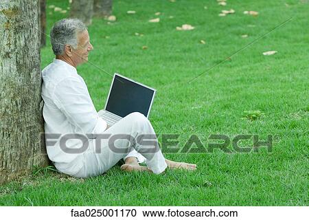 Mature man sitting in grass, leaning against tree, using laptop computer View Large Photo Image Stock Image - Mature man sitting in grass, leaning against tree, using laptop computer. Fotosearch