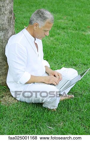 Mature man sitting in grass, leaning against tree, using laptop computer View Large Photo Image Stock Photo - Mature man sitting in grass, leaning against tree, using laptop computer. Fotosearch