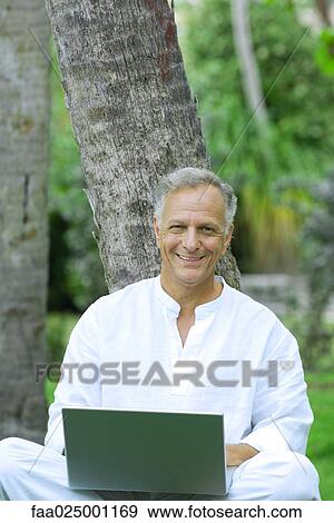 Mature man sitting, leaning against tree, using laptop computer, smiling at camera View Large Photo Image Stock Photo - Mature man sitting, leaning against tree, using laptop computer, smiling at camera. Fotosearch