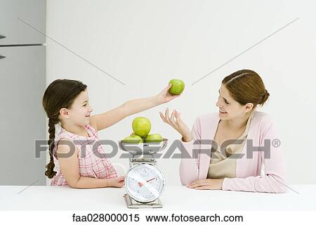 Stock Photography - Mother and daughter weighing apples on scale, smiling at each other, girl holding up one apple. Fotosearch