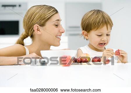 Stock Photograph - Mother and son in kitchen with assorted ingredients, boy holding grape, both smiling. Fotosearch