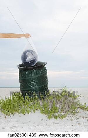 Stock Photo - Person holding globe in plastic bag over garbage can, cropped view. Fotosearch