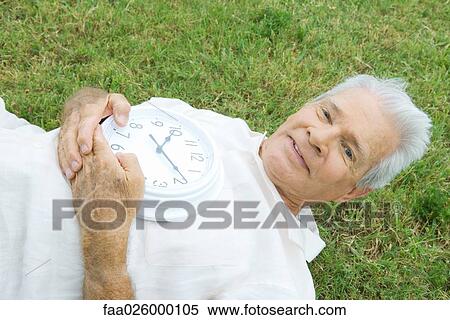 Senior man lying on grass, holding clock, smiling at camera View Large Photo Image Stock Photography - Senior man lying on grass, holding clock, smiling at camera. Fotosearch